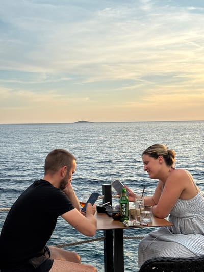 Two people, a man and a woman, are seated at a table by the sea at sunset. The man, on the left, wears a black t-shirt and shorts and looks down at his phone. The woman, on the right, wears a striped dress and is also looking at her phone, smiling. On the table are drinks, cigarettes, and a small bowl of nuts. In the background, the sea stretches to the horizon, with a small island visible in the distance. The sky is a pale blue with wispy clouds, tinged with the warm colors of the setting sun. This scene is likely located in Primošten, Croatia, a coastal town known for its picturesque sunsets over the Adriatic Sea.