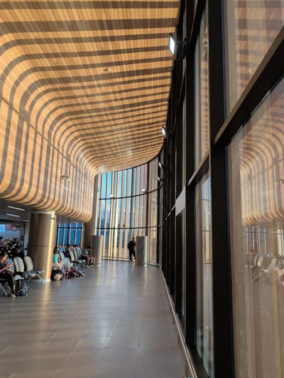 The image depicts the interior of a modern airport terminal in Tambon Hua Toei, Thailand. Sunlight streams through a large, curved glass facade, casting striped shadows across a wooden ceiling and floor. Several people are seated on benches, some looking at their phones. A person stands near the windows. The terminal appears clean and well-lit, with a polished concrete floor and metal chairs. The architecture is contemporary, featuring curved lines and natural materials like wood and glass. The overall impression is one of a spacious and bright waiting area, likely at an airport or similar transportation hub.