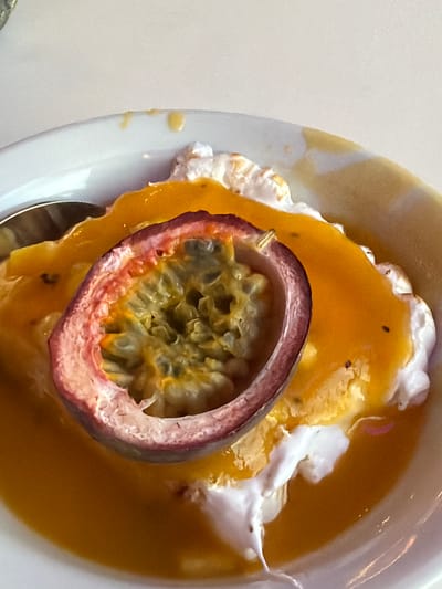 This is a close-up, overhead shot of a dessert in a white bowl. The dessert appears to be a passion fruit parfait, with a thick, orange passion fruit sauce covering a layer of white whipped cream or yogurt, and topped with a halved passion fruit. The passion fruit, with its purple rind and green, seed-filled interior, is prominently displayed. The bowl is white and has a slight rim. A silver spoon is visible on the left side of the bowl, partially submerged in the dessert. The background is a plain, light-colored surface, likely a table. The lighting is bright, casting soft shadows. This dessert was photographed on July 16, 2023, at 1:22 PM in Pierrelatte, France.