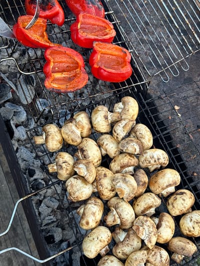 The image shows red bell peppers and whole button mushrooms grilling on a charcoal grill. The grill sits above grey charcoal. A fork is inserted into one of the pepper halves. The location is not discernable from the image.