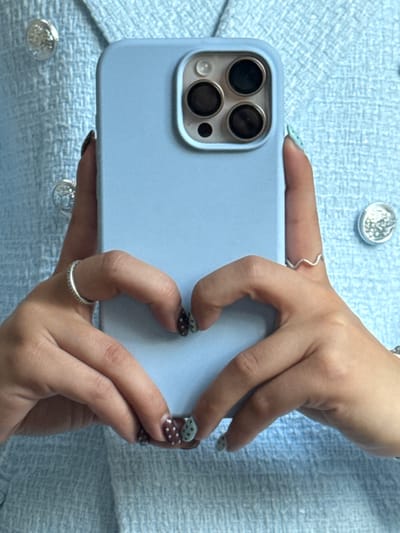 A person is holding a light blue smartphone with a visible camera module featuring three large lenses and a smaller sensor or flash. The phone is held horizontally, with the camera facing left. The person's hands, adorned with rings and patterned nail polish, are gripping the edges of the phone. The person is wearing a light blue tweed-like jacket or top with decorative silver buttons. The background is the same light blue tweed-like fabric. There is no geographical information about Bruchköbel, Germany in the image.