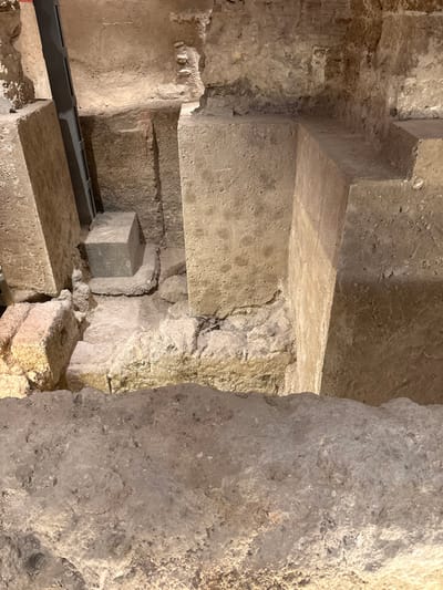 The image displays an archaeological excavation site, likely located in the city of Cádiz, Spain, featuring ancient stone and earthen structures. The perspective is from an elevated viewpoint, looking down into a subterranean or deeply excavated area.

In the foreground, an uneven, light-colored earthen or concrete edge defines the immediate viewing platform. Below this, the scene reveals several prominent, roughly hewn rectangular stone blocks functioning as pillars or foundational elements. On the left, a tall, light-brown textured stone pillar stands, with a dark metal support beam or structure partially visible behind it. To the right of this pillar, a smaller, dark grey cube-shaped block rests on a raised, irregular stone base. Further to the right, another substantial light-brown stone pillar forms a corner or wall segment. The exposed ground within the excavation is uneven, consisting of layered earth and irregular stone formations, including some reddish-toned blocks visible on the lower left. The environment is characterized by exposed ancient building materials and earth, illuminated by artificial light. No people or active elements are present in the static scene.