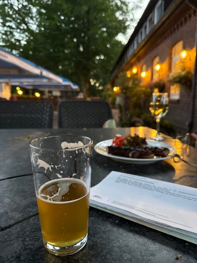 This is a close-up, low-angle shot of a partially full glass of beer on a dark, textured table, likely outdoors. The beer is a pale golden color with some foam clinging to the sides of the glass. The glass has a logo with some text and imagery, though it is partially obscured by the beer and foam. In the background, out of focus, are several elements that provide context.

To the left of the beer glass, there's a plate with some dark food, possibly meat, and a small garnish of lettuce and a red item, perhaps a piece of tomato. Behind the plate, a blurry image of a building with warm lights emanating from its windows suggests a restaurant or pub. Strings of fairy lights are also visible, adding to the ambient lighting. The scene appears to be set in the evening or late afternoon, as indicated by the warm, artificial lights.

Further in the background, a blurry expanse of green foliage suggests trees or a garden. To the right of the beer glass, a white document or menu is partially visible, with some German text printed on it. The text visible on the document mentions "Bundesdruckerei" (Federal Printing Office) and details opening hours, stating "Montag - Freitag von 09:00 Uhr bis 16:00" (Monday - Friday from 9:00 AM to 4:00 PM) and refers to "gesetzliche Feiertage" (public holidays).

The overall impression is a relaxed, outdoor dining or drinking setting, possibly at a German establishment, with a focus on the beer and the food. The blurred background contributes to a sense of depth and atmosphere, highlighting the primary subjects.
