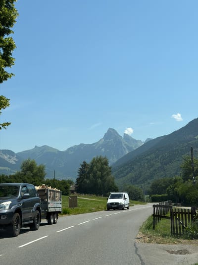 This image depicts a scenic mountain landscape with a road running through it. The foreground shows a gray pickup truck towing a trailer filled with firewood. Further down the road, a white van is driving towards the viewer. The road is paved and has white lane markings. To the left of the road, there is a grassy area with a wooden fence. Beyond the road and grassy area, a dense forest covers the lower slopes of the mountains. The mountains themselves are rugged and layered, fading into the distance. The sky is a clear, bright blue with a few small white clouds. The sun appears to be high in the sky, suggesting it is midday. The overall impression is one of a beautiful, natural setting on a clear day. There is no visible text in the image.