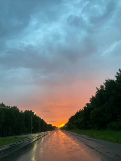 The image depicts a wet asphalt road stretching into the distance under a dramatic twilight sky. The sky transitions from a cool blue-gray at the top to a warm gradient of pink and orange near the horizon, where the setting sun is partially obscured by clouds. Dense green trees line both sides of the road, creating a tunnel effect. The road surface is reflective, with puddles of water mirroring the sky and the lights from street lamps positioned along the left side of the road. A single car with illuminated headlights is visible driving away from the viewer, further down the road. The scene appears to be a rural or suburban highway, possibly in the vicinity of Nurykeevo, Russia, given the information provided.