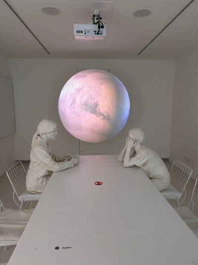 A shot of a room at Casa de los Volcanes in Haría, Spain. The image appears to be rotated. A large illuminated sphere resembling the planet Mars hangs in the center of the room. A white projector is mounted on the wall. Two white mannequin figures are positioned on a white table with wire mesh shelving underneath. One figure rests its face in its hands, while the other reaches forward with one hand holding a small object.