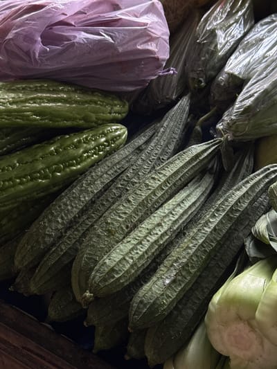 A close-up, overhead view captures a collection of fresh vegetables for sale, likely at a market. Dominating the frame are several ribbed, green gourds, some smooth and elongated, others with a more textured surface. To the left, partially obscured by the gourds, are two pale, waxy gourds. A light purple plastic bag lies at the top of the image, and further back, several vegetables are wrapped in clear plastic. The scene is dimly lit, with shadows accentuating the textures of the produce. The location is a market stall in Karangsambung, Indonesia.