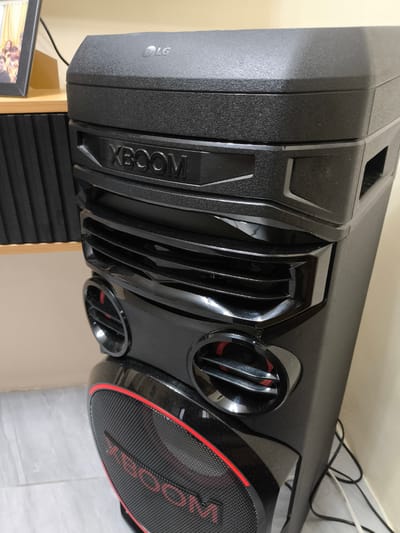 This is a close-up, eye-level shot of an LG XBOOM speaker, a large, black, vertical audio device. The speaker is positioned on the right side of the frame, taking up most of the image. It has a textured, matte black finish on its top and side panels. Prominently displayed on the front of the speaker, below the top panel, is the "XBOOM" logo in raised, glossy black lettering. Below this, there is a grille-like design with horizontal slats, also in glossy black. Further down, two circular speaker vents with red accents are visible. The bottom section of the speaker features a large, mesh-covered speaker with a red trim and the "XBOOM" logo in red lettering across the mesh.

To the left of the speaker, a portion of a wooden shelf is visible, holding a framed photograph of several people. The background consists of a plain, light-colored wall. The lighting appears to be indoor, likely ambient room light, suggesting a daytime setting within a home or establishment. There are no clear indications of weather conditions. The overall impression is one of a functional audio device in a domestic or retail environment. The presence of a framed family photo hints at a personal space. No specific activity is evident, but the object's purpose implies sound or music would be played. Visible text includes "LG" and "XBOOM". The location context is Imus, Philippines.