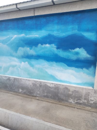The image displays a concrete wall in Chighirik, Uzbekistan, featuring a large mural. The mural, covering the upper section of the wall, depicts a landscape of blue mountains partially obscured by white and light-blue cloud formations, transitioning from darker blue hues at the top to lighter shades towards the bottom. Below the mural, the wall consists of exposed, unpainted concrete, exhibiting textured surfaces and areas of discoloration. Above the mural, a light gray concrete structure with dark details, possibly an eave or roofline, is visible. In the foreground, a concrete ground surface extends horizontally, featuring a shallow, linear concrete channel running parallel to the wall. No individuals, discernible actions, or direct interactions are visible within the frame.