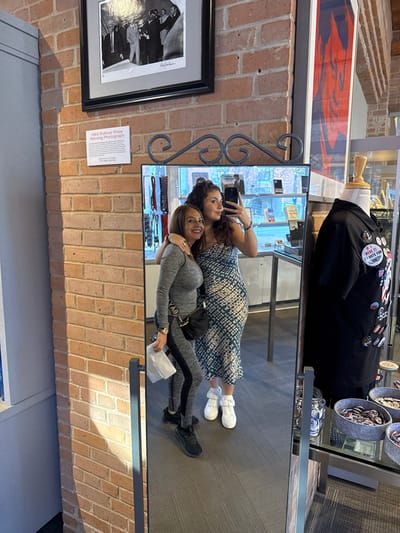 Two women are reflected in a mirror. One woman wears a printed dress and white sneakers, holding a phone in her hand. The other woman wears a gray tracksuit and black sneakers, standing slightly behind the first woman with an arm around her. The mirror hangs on a brick wall next to framed photographs. The reflection shows the floor and a glass display case containing items like buttons and clothing.