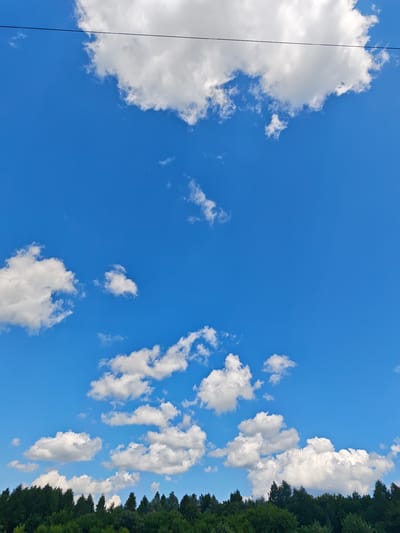 The image shows a bright blue sky filled with scattered white cumulus clouds. In the foreground, a dense line of green trees forms a forest canopy against the horizon. The photo appears to have been taken outdoors on a clear day. There are no people or animals visible in the image. A thin black wire is visible stretching across the upper portion of the frame, suggesting the presence of a power line or telephone line. The overall scene depicts a natural landscape with a touch of human infrastructure. The location is identified as being in the city of Kozhlasola, Russia.
