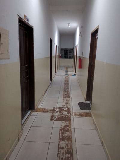 The image shows a long, narrow, brightly lit interior hallway with white walls and a beige stripe along the lower portion. The floor is tiled with large, light-colored square tiles, some of which are stained and scuffed, particularly in a wide central stripe extending down the hallway. There are several dark wooden doors on both sides of the hallway, leading into rooms that are not visible. A sign with the word "ZEBRA" is visible above the first door on the left. Further down the hallway on the left, a metal-grated window or opening is visible. On the right side of the hallway, there is a red fire extinguisher mounted on the wall. The hallway extends into the distance, where it appears to curve slightly. The lighting is a combination of overhead fluorescent lights and light coming from the end of the hallway. The overall impression is one of a functional, institutional space. The location is identified as Cáceres, Brazil.