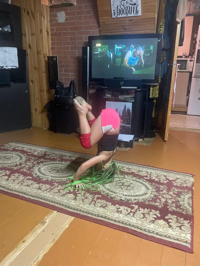 In a dimly lit room, a young person with vibrant green braided hair is positioned in a prone, almost acrobatic pose on a patterned rug. They are wearing a dark tank top and bright pink shorts with white trim. Their bare feet, clad in what appear to be ballet slippers or similar soft shoes, are propped up on a black bag.

The room has a rustic feel, with wooden plank walls and a brick-patterned backdrop behind a television set. The television screen displays an image of a man in a blue tank top, seemingly engaged in some form of exercise or activity. A shelf above the television holds various items, including what looks like a speaker and some fabric with a tiger print pattern. A small white sign with decorative text is visible on the wooden wall to the left of the television.

The overall impression is one of a casual, perhaps personal space, possibly a living room or a dedicated practice area. The lighting suggests it could be evening or a naturally dark day. The focus is on the individual in the foreground, whose pose and unique hair color are the most striking elements. The presence of exercise equipment (the television program and the black bag) hints at a focus on physical activity.
