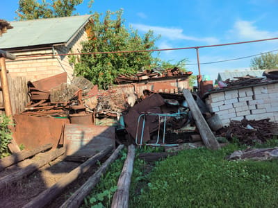 The image depicts a cluttered outdoor scene in Алькино, Russia, likely a yard or storage area. A large pile of scrap metal and other discarded items is the central subject. The materials include rusted barrels, sheets of metal, wire cages, wooden planks, and what appears to be parts of a bicycle or cart. In the background, a light-colored brick building with a green metal roof is visible, partially obscured by a lush green tree laden with red berries. Further in the distance, a white brick wall with more piled-up debris is present. The ground is covered with dry earth and patches of green grass. Several weathered wooden logs are placed horizontally on the ground in the foreground.
The time of day appears to be late afternoon or early evening, given the strong shadows cast by the objects and the warm sunlight. The weather is clear and sunny, with a bright blue sky and a few white clouds. There are no people visible in the image. The overall impression is one of disarray and neglect, suggesting a place where items are stored or accumulated for later disposal or recycling. No text is visible in the image.