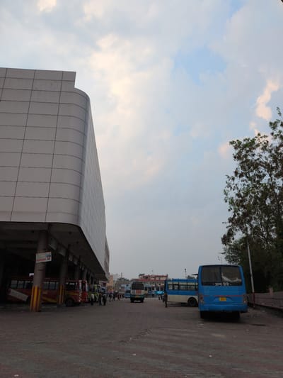The image depicts a bus station in Una, India. In the foreground, a blue bus is parked facing away from the viewer, with visible Indian license plates and Hindi script on its rear window. To the left, a modern, multi-story building with a curved facade and white paneling dominates the scene. Underneath the overhang of this building, several other buses are parked, including a red and yellow bus and a green bus. A few people are visible standing near the buses, some appearing to be waiting or interacting. The ground is paved with rough asphalt. In the background, other buildings and trees are visible, suggesting an urban environment. The sky is cloudy with patches of blue and pale yellow light, indicating it might be early morning or late afternoon. The overall scene is that of a busy public transportation hub.