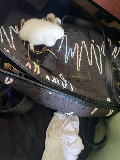 The image shows a black handbag with colorful wavy patterns. A fluffy white keychain is attached to the bag. The bag is placed on top of dark clothing, with a white garment partially visible. The scene is indoors. There is no information to suggest that this picture was taken in Sabo, Nigeria.