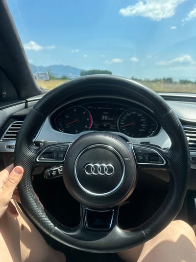 A first-person perspective shot shows a person's hand gripping the steering wheel of a car. The steering wheel is black leather with red stitching and the Audi logo is prominently displayed in the center. The dashboard and instruments are visible, showing a speed of 39 km/h. The car is driving on a sunny day, with a clear blue sky and some clouds visible through the windshield. The background shows a rural landscape with fields and distant mountains. The scene appears to be from a road in or near Ovoshtnik, Bulgaria, given the context provided. The overall impression is that of a leisurely drive on a warm day.