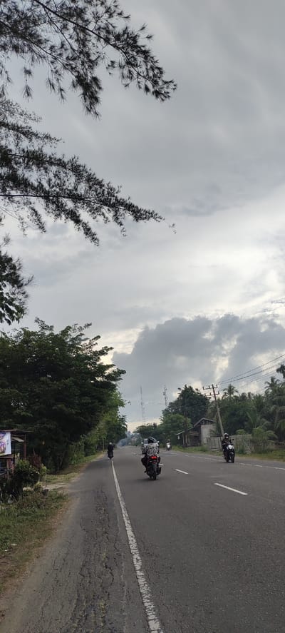 The image depicts a two-lane asphalt road in Aluekeujruen, Indonesia, under a cloudy sky. Several motorcycles are in motion on the road, with riders wearing helmets. On the left side of the road, there are trees and some vegetation, along with a small structure that appears to be a roadside stall with a banner. On the right side, there are more trees, some buildings, and utility poles. The road surface shows signs of wear and tear, with cracks and uneven patches. The overall scene suggests a rural or semi-rural setting on a day with overcast weather.