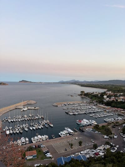 The image presents a high-angle view of a harbor filled with numerous sailboats and motorboats docked in calm water. The harbor is bordered by a town with buildings clustered along the coastline, surrounded by greenery. In the distance, a peninsula with a hill extends into the sea, and a range of mountains forms the backdrop. The sky transitions from a pale blue at the top to softer pinks and oranges near the horizon, suggesting either sunrise or sunset. The scene is static, with no visible human activity. The overall impression is one of a peaceful coastal town and its bustling marina.