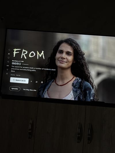 The image shows a TV screen displaying the title screen of the TV show "From." A woman with curly hair and a red necklace is visible in the background image of the screen. Below the title, text describes the show's plot and indicates it is a 4-season series that airs on HBO, and is labeled with the drama and horror genres. A "Watch S4 E3" button is highlighted. The TV is situated next to a dark wood-look dresser with metal handles. The location and city of Selanovtsi, Bulgaria is not visible.
