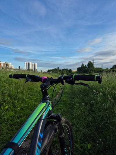 The image shows a first-person perspective of a person sitting on a bicycle on a dirt path in a grassy field. The bicycle is black with green and blue accents. The handlebars are visible, with grips and brake levers. In the background, there are several multi-story apartment buildings, some with white facades and others with lighter colored facades and dark windows. There is also a line of trees and what appear to be power lines in the distance. The sky is blue with wispy white clouds and a sliver of a crescent moon is visible. The location appears to be the outskirts of Yanino-2, Russia.