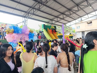 The image depicts a crowded outdoor event in Timaná, Colombia, under a metal-framed roof structure. A large group of people, including children and adults, are gathered. Features include a decorated backdrop, balloon arrangements, and an inflatable slide. The background shows buildings and vegetation.