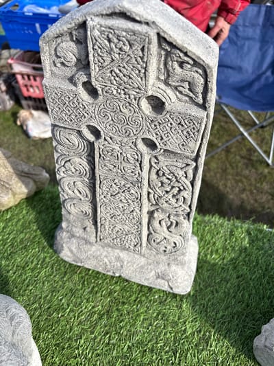 The image shows a grey, intricately carved Celtic cross lying on artificial grass. The cross is the main subject and features detailed knotwork patterns and possibly animal motifs. A portion of a person's red-sleeved arm and hand are visible in the upper left, along with a blue plastic crate and part of a metal folding chair. A partially visible stone is near the right side. The background is blurred, but suggests an outdoor setting with a mix of natural and artificial elements. The image was taken in Chirk, United Kingdom.