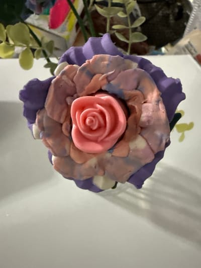 A close-up, eye-level shot features a flower sculpture made of modeling clay, with delicate purple petals forming the outer layer and a bright pink rosebud at its center. The inner petals are a mix of pastel colors, including pink, white, and blue, creating a marbled effect. The sculpture is placed on a white surface, and to the left, a portion of a decorative artificial plant with pale green leaves is visible. The background is blurred, hinting at more objects, including what appears to be a dark, rocky object. The lighting casts soft shadows on the white surface, suggesting an indoor setting. The image is taken in Cartersville, United States.