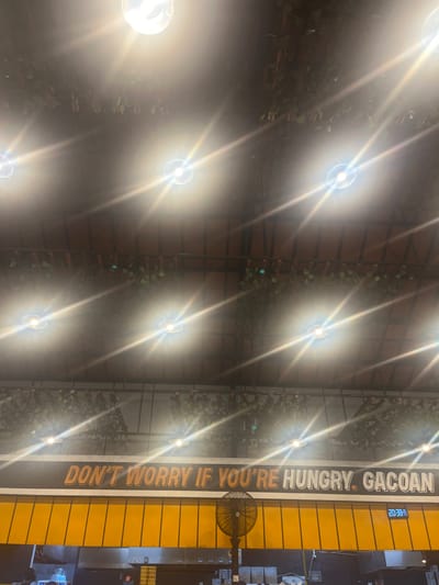 The image captures an interior view of a restaurant or eatery, likely in Binong, Indonesia, as indicated by the sign. The scene is illuminated by bright overhead lights, creating strong glare and lens flares that obscure some details. A prominent sign on the wall reads "DON'T WORRY IF YOU'RE HUNGRY. GACOAN", suggesting it is an advertisement for the restaurant chain Gacoan. Below the sign, a bright yellow awning covers the top of what appears to be a service counter or kitchen area. A black oscillating fan is positioned to the left of the awning. Various kitchen equipment and possibly some patrons are dimly visible in the lower part of the image, beneath the counter. The ceiling features wooden beams and decorative artificial greenery.