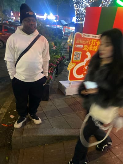 The image depicts an outdoor night scene in Tian'gongdian Subdistrict, China, featuring two individuals on a paved pedestrian area illuminated by artificial lights.
In the foreground to the left, a dark-skinned adult male stands, facing slightly right. He wears a black beanie, a white long-sleeve hooded sweatshirt with black stripes visible on the sleeves, black sweatpants, and light-colored slip-on shoes with black elements. A black strap crosses his torso from his right shoulder to his left hip. His hands are in his pockets, and his gaze is directed towards the right side of the frame.
To the right, an adult female is captured in motion blur, walking from right to left. She has long dark hair and is wearing a dark, possibly black, textured jacket or coat, dark pants, and dark shoes with white soles. She carries a white shoulder bag with a thick white strap and holds a white object, possibly a mobile phone, in her right hand.
Between the two individuals is a rectangular planter containing pink flowers and green foliage. To the right of the standing male and partially obscured by the moving female is a large vertical promotional sign with a red background. The sign features Chinese characters, including "大众点评" (Dianping) and "紫微路" (Ziwei Road), a QR code, dates "23.12.9 - 24.3.3", and graphic elements of a winding road and a cartoon character.
The background reveals a brightly lit urban environment at night. To the far left, a red vehicle is partially visible. Further back, numerous bright, blurred lights suggest shops or market stalls, some with blue canopies. To the far right, a tree wrapped in numerous small white lights is visible. The ground is covered with square grey paving tiles.