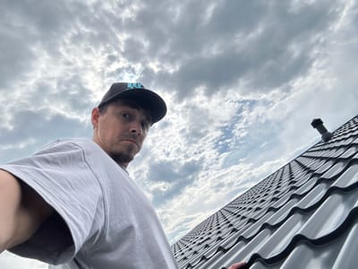 The image, taken in Rassokha, Russia, shows a low-angle, close-up, selfie-style shot of a man on a rooftop. He is wearing a grey t-shirt and a dark baseball cap with a blue logo. His face is partially visible, looking towards the camera with a neutral expression. The background consists of a cloudy sky and a section of a dark, wavy metal roof, with a pipe or vent visible on the roof. The lighting suggests it is daytime, possibly overcast.