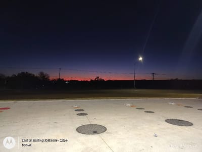 A pre-dawn landscape displays a concrete area with circular manhole covers and square access points. Beyond, a thin strip of red-orange horizon sits below a dark blue sky. Silhouetted trees and utility poles break the horizon line. A street lamp glows on the right, casting light and a long beam into the sky. A timestamp and watermark are in the lower corners of the image.