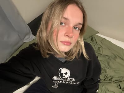 A person with fair skin and blonde hair is lying down on a bed with green and blue bedsheets. They are wearing a black hooded sweatshirt with a white graphic on the front. The person has freckles, and is wearing a nose ring. They are looking up and to the side, with their head tilted.