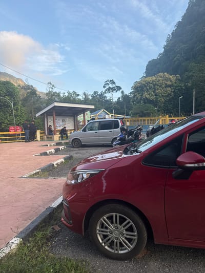 In Wang Kelian, Malaysia, a red car is parked in the foreground, angled towards the right. Behind it, a silver van is parked. Further back, a small open-air shelter with a white roof and lattice sides is visible. Three men are standing outside the shelter, and two more men are seated inside. Several motorcycles are parked near the van. The area appears to be a parking lot or a rest stop. In the background, a large, tree-covered mountain looms under a partly cloudy sky. A yellow fence runs along the back of the scene.