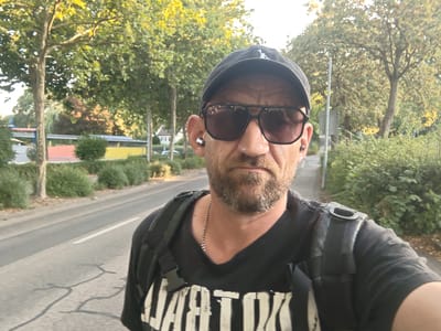 A selfie taken by a man with a beard, wearing a black baseball cap, sunglasses, a black t-shirt with white lettering, a silver chain necklace, and a black backpack with straps visible. He is wearing wireless earbuds in both ears. The man is standing outdoors on a paved walkway next to a road. Trees with green and yellow foliage are visible behind him. In the background, there are buildings with red and yellow roofs, and dense green bushes. The lighting suggests it is daytime, with sunlight filtering through the trees. The environment appears to be a suburban or semi-rural area. The location is identified as Nordheim, Germany.