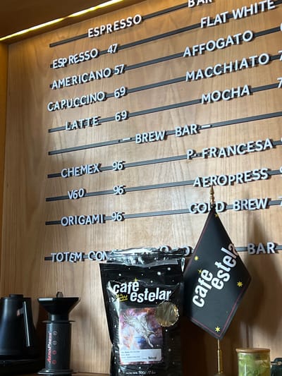 The image shows a coffee menu board in a store selling Café Estelar coffee, likely located in Mazatlán, Mexico. The menu lists different coffee drinks and their prices, including espresso, americano, cappuccino, latte, Chemex, and Aeropress. There is also a bag of Café Estelar coffee beans next to an Aeropress brewing device. The board is made of wood, with white lettering on black rods. The text is printed in both English and Spanish. The overall setting appears to be a coffee shop.