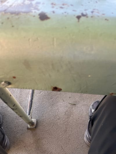 The scene depicts a low-angle view over a horizontal, light green-painted metal surface, exhibiting significant wear, numerous irregular brown rust spots, and some chipped paint. Faint vertical lines are visible on this surface. Below it, two metal support poles are visible; the left pole is light gray and connected to a square metal base plate resting on a light gray, textured concrete ground. The right pole is darker and partially obscured. On the concrete surface, a single thin, green leaf or blade of grass is present. The lower legs and feet of an individual are visible at the bottom edges of the frame, indicating the camera's perspective from a person's viewpoint. The person wears dark pants and dark athletic shoes with white soles and lighter accents. The location is Krushovo, Bulgaria.
