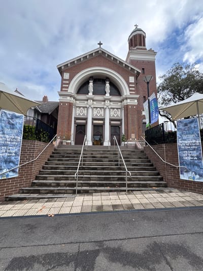 This image captures the entrance to a brick church, likely a Catholic parish, given the "Our Lady of Dolours" text on a banner and the religious iconography. The church features classical architectural elements, including a grand arched entrance, columns, and statues. A prominent bell tower with a cross tops the building.

The scene is set outdoors with a slightly overcast sky, suggesting a daytime setting. A set of wide, stone stairs with metal handrails leads up to the church entrance. Two banners are visible: one at the top of the stairs, featuring images and text that reads "Bringing the light of Christ to the city, We Love, We Grow, We Serve," and another smaller banner on a pole near the bottom of the stairs that says "WELCOME" and "Benvenuti."

The overall impression is one of a welcoming and established place of worship. There are no people visible in the image, focusing the attention on the architecture and the welcoming messages. The presence of fallen leaves on the stairs and pavement hints at an autumn or early winter season. The location context provided is Roseville Chase, Australia, which helps to place the church in a specific geographical setting.
