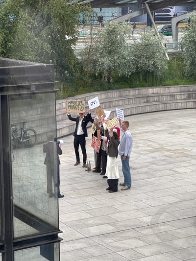 The image, taken from a slightly elevated viewpoint, captures an outdoor urban scene in Tårnby, Denmark, featuring a small protest or demonstration. A group of approximately five individuals stands on light-colored paved ground, holding protest signs. Behind them, wide concrete stairs ascend, and to their left, lush green trees and foliage are visible. Modern glass-fronted buildings and reflections of a white Mercedes car and a bicycle can be seen in the background and foreground, indicating a contemporary urban environment.

The protesters are holding signs with various messages. One prominent sign, held by a person in a dark suit, reads "HANDS OFF MY PRIVATE JET" and an adjacent sign says "OV!". Another sign, further back in the group, displays the Danish text "AFGIFTER ER KUN FOR ALMINDELIGE DANSKERE", which translates to "TAXES ARE ONLY FOR ORDINARY DANES". A red sign is also visible, stating "ELITEN SIGER FRA", meaning "THE ELITE SAYS NO" or "THE ELITE STEPS BACK". The demonstrators appear to be engaged in a focused, peaceful protest.

The setting suggests an urban plaza or public space. The weather appears to be overcast daytime, with soft lighting and no harsh shadows. A glass barrier or railing is prominently featured in the immediate foreground, showing reflections of the surroundings.