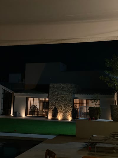 The image presents an outdoor scene at night, likely in Santa Cruz Tehuixpango, Mexico, given the prompt. A modern building with a stone facade and multiple windows is partially visible. Some of the windows are illuminated, revealing interior details such as furniture. An expanse of green lawn extends alongside the building. To the side of the building, there appears to be a partially obscured white canvas or screen. A dark, possibly treed area is in the background. The overall impression is that of an upscale residence with outdoor lighting. A human figure is partially visible in the distance.