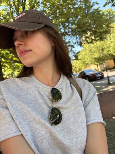 The image is an upward-angled selfie of a young woman in an outdoor setting. She is wearing a gray t-shirt, a brown baseball cap, and has sunglasses resting on her shirt. Her dark hair is visible beneath the cap. The background includes trees and a glimpse of a street with a car. While the exact location in Siófok, Hungary, cannot be determined from the visual data alone, the presence of trees and a street suggests a park or roadside area within the city.