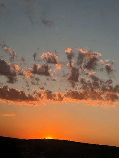 The image depicts a sunset over a rural landscape. The sky is a gradient of soft oranges and yellows, with a few scattered clouds tinged with pink and brown. The sun, a bright orange orb, is low on the horizon, partially obscured by a dark, silhouetted landmass on the right side of the frame. This landmass appears to be a hill or a ridge, with faint outlines of trees or buildings visible against the sky. The left side of the image shows a lighter blue sky, with more wispy clouds. The overall impression is one of tranquility and the end of a day. The location context of Hrabovets, Ukraine, suggests a natural, perhaps agricultural or forested, setting. The time of day is clearly sunset. The weather appears clear, allowing for a vibrant sunset display. There is no visible text in the image.
