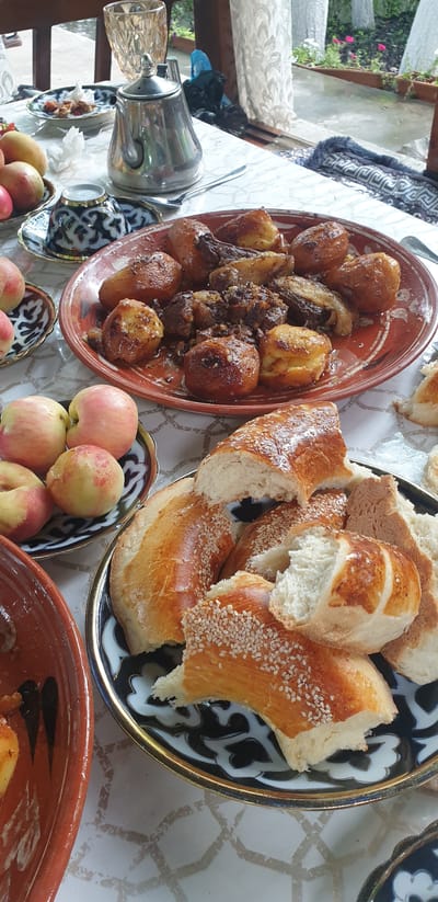 A table is set with a meal in Avval', Uzbekistan, possibly outdoors or in a room with a view of trees and foliage. The table is covered with a white cloth. In the center is a large, reddish-brown ceramic platter filled with roasted meat and potatoes. To the right of this platter is a dark-colored ceramic plate piled with pieces of sesame seed-crusted bread. In the foreground, on the left, is a reddish-brown ceramic bowl containing food. To the left of the meat and potato platter is a small, dark-colored dish with white elements, and to its left are a few ripe apples on a dark-patterned plate. Behind the apples, a silver teapot sits next to a small, dark-patterned cup and saucer. A tall, faceted glass is visible in the background. The scene suggests a meal in progress or just served.