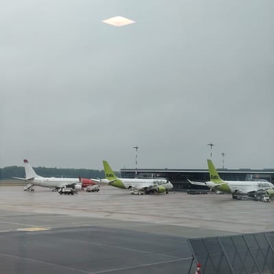 The image displays an outdoor airport scene at Riga International Airport, near Skulte, Latvia, under an overcast sky. Three commercial passenger aircraft are parked on a grey asphalt and concrete apron.

On the far left, a white Boeing 737 aircraft is visible, featuring a red tail with "norwegian.com" text and a partial registration of "LN-NOK" on the fuselage. A portable passenger staircase is positioned at its front door, and a white ground service vehicle is located beneath its fuselage.

In the center, a white Airbus A220 aircraft, registered YL-CSG, is prominently displayed. It has a distinctive lime green tail marked "Baltic" and matching lime green engine nacelles with "airBaltic" branding. Multiple ground support vehicles, including a mobile passenger stair unit, are actively engaged around the aircraft's undercarriage and fuselage.

To the right, partially visible, is another airBaltic Airbus A220 aircraft, identified by its similar lime green tail with "Baltic" text and "airBaltic" branding. Ground support equipment is also visible around this aircraft.

In the background, behind the two airBaltic aircraft, a modern airport terminal building with a dark-colored facade and extensive glass windows is present. Between the aircraft and the terminal, several tall, black and white striped utility poles equipped with instruments are visible.

The tarmac surface appears damp or wet in various areas, suggesting recent precipitation or high humidity. Yellow and white painted markings are present on the apron surface. Small, indistinct figures of ground crew are discernible near the central airBaltic aircraft.

In the immediate foreground, a dark grey metal fence with a diamond grid pattern partially obstructs the lower right portion of the view. A square, illuminated light fixture is reflected in the upper central portion of the image, indicating the photograph was taken from inside a building through a window.