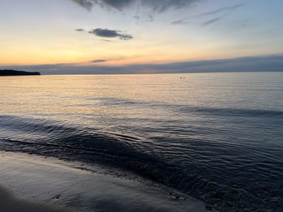 The image shows a serene coastal scene at dusk in Dronningmølle, Denmark. The sky is a gradient of soft blues, greys, and warm yellows, with a few scattered clouds. The sea is calm, with gentle waves lapping at the sandy shore. The water reflects the colors of the sunset, creating a tranquil atmosphere. In the distance, a faint silhouette of land is visible on the horizon, possibly Mølle strand. There are a few dark figures in the water, appearing to be people, some partially submerged. The foreground consists of wet sand with small pebbles and ripples from the receding waves.