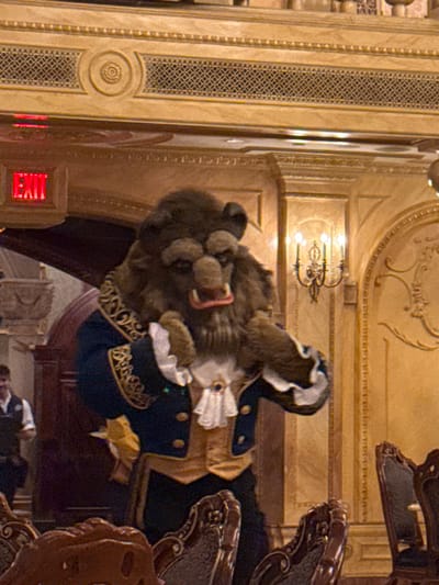This image depicts an indoor scene within the Be Our Guest Restaurant, located in Magic Kingdom at Walt Disney World Resort, Bay Lake, Florida, United States.

A costumed performer portraying the Beast character from Disney's "Beauty and the Beast" stands centrally. The performer wears a brown fur costume featuring a pronounced snout, fangs, and an open mouth with visible pink gums. The costume includes a dark blue tailcoat with elaborate gold embroidery on the lapels and cuffs, a tan-colored waistcoat, a white ruffled jabot, and white ruffled cuffs. The performer's left arm is slightly bent, with the hand raised to chest height.

The background features opulent architecture consistent with a grand dining hall. Walls are a light golden-brown, adorned with intricate decorative moldings, fluted pilasters, and stylized bas-relief patterns. A dark metal wall sconce with three illuminated candle-like bulbs is mounted on the wall to the right. In the upper left, a red "EXIT" sign with white lettering is illuminated above a dark archway. Partially visible in the lower left background, a male individual wearing a white shirt and dark vest suggests a staff member. The immediate foreground shows the dark, ornate backs of several dining chairs, indicating the presence of tables. The scene is illuminated by ambient artificial light.