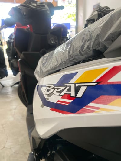 A close-up, side-view, eye-level shot shows the white body panel of a Honda Beat scooter, prominently displaying the "BEAT" logo in black and white, outlined in blue and red. The scooter is parked indoors, likely at a dealership, as indicated by the plastic wrap on the seat and the presence of other vehicles in the background. The scooter's design features a red, white, blue, and yellow graphic pattern. The floor appears to be concrete, with some scuff marks. The overall lighting suggests an indoor, well-lit environment. The image captures a section of the scooter's front fairing and part of its seat. The location is identified as San Jose del Monte, Philippines.