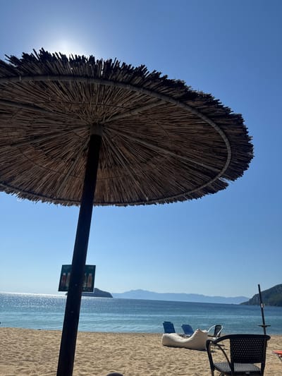 This image captures a sunny beach scene in Nea Iraklitsa, Greece. In the foreground, a large thatched umbrella, casting a shadow, dominates the view. A wooden pole supports the umbrella and has a small sign attached to it, displaying what appear to be two bottles of beverages. The sandy beach stretches out towards the turquoise sea. In the midground, several pieces of beach furniture are visible: a large white beanbag chair and two blue lounge chairs are arranged on the sand. Further out, the calm sea reflects the bright sunlight, and a hazy silhouette of distant mountains and islands can be seen on the horizon. The sky is clear and a vibrant blue.