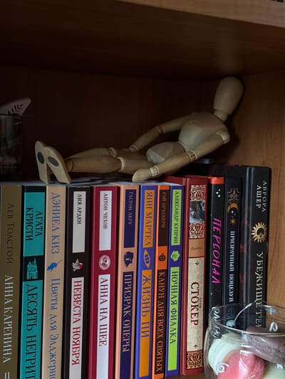 The image shows a wooden mannequin lying on its back on a shelf in a dimly lit wooden bookcase. The mannequin is positioned such that its head and torso are towards the back of the shelf, and its legs are extended forward, resting on the spines of several books. The mannequin is made of light-colored wood and has articulated joints.
Below the mannequin, a row of books is visible, arranged vertically. The spines of the books are colorful and feature text in Russian. Some of the visible titles include "Лев Толстой", "Анна Каренина", "Агата Кристи", "Дэниел Киз", "Цветы для Элджернона", "Невеста ноября", "Анна на шее", "Гастон Леру", "Призрак Оперы", "ЯНН МАРТЕА", "КАНУН ДНЯ ВСЕХ СВЯТЫХ", "Ночная Фиалка", "СТОКЕР", "Персонал", "Призрачный поцелуй", "Аврора Ашер", and "Убежище". A glass jar containing several colorful objects, possibly soaps or macarons, is positioned in front of the books on the lower shelf, partially obscuring some of the book spines. The environment appears to be an indoor setting, likely a home or a library, with wooden shelving. There are no other people or living beings visible in the image. The lighting is soft and warm, casting shadows that highlight the textures of the wood and the book covers. The overall impression is one of a quiet, staged scene.