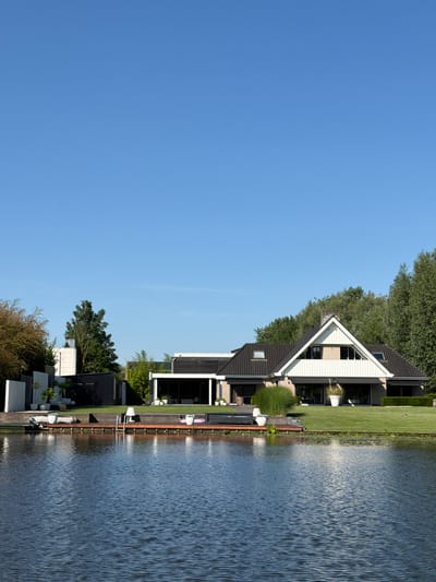 The image depicts a waterside property in Acquoy, Netherlands. A modern, multi-level house with a dark tiled roof and large windows is situated on a manicured lawn behind a wooden deck. The deck extends to the edge of a calm body of water. On the deck, there are several white planters, a ladder leading into the water, and a white lamp. Beyond the house, mature trees form a dense green backdrop against a clear blue sky. In the foreground, the dark blue water shows ripples, suggesting a gentle breeze. The overall scene conveys a sense of tranquility and modern living.